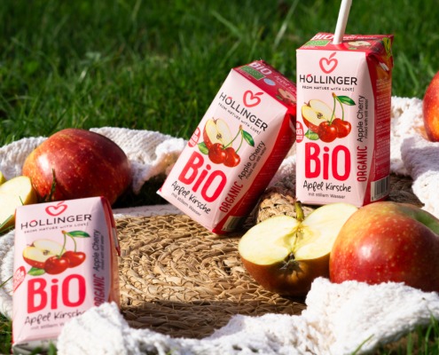 Höllinger Bio Apfel-Kirsch-Schulsaftpackungen und frische Äpfel auf einer Picknickdecke im Gras.